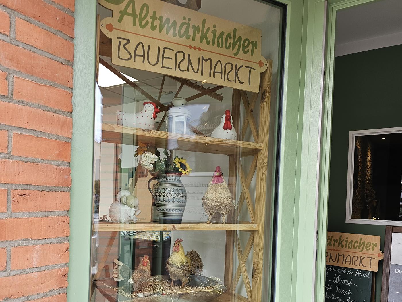 Das Schaufenster neben der grünen Ladentür, darin ein Holzregal mit Keramikhühnern und Krügen, darüber das Holzschild Altmärkischer Bauernmarkt.