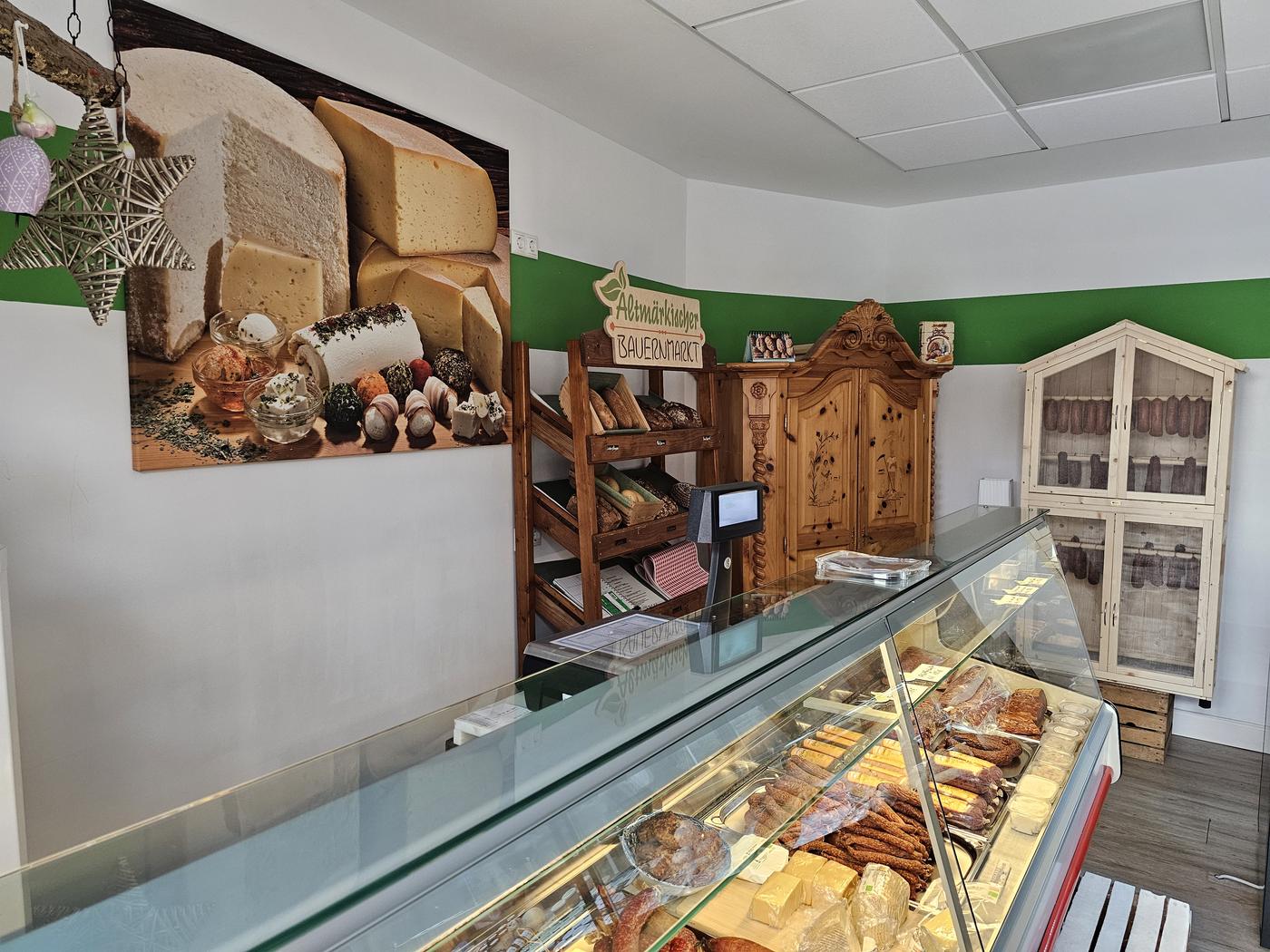 Die Verkaufstheke mit Wurst und Käse, dahinter das Brotregal, ein Bauernschrank und der Wurstschrank aus hellem Holz.