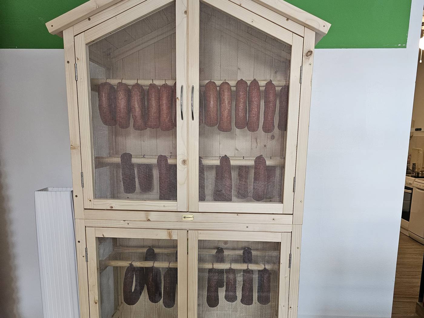 Ein hoher Schrank aus hellem Holz mit Gitterfenstern, darin hängen Dauerwürste und Wurstringe.