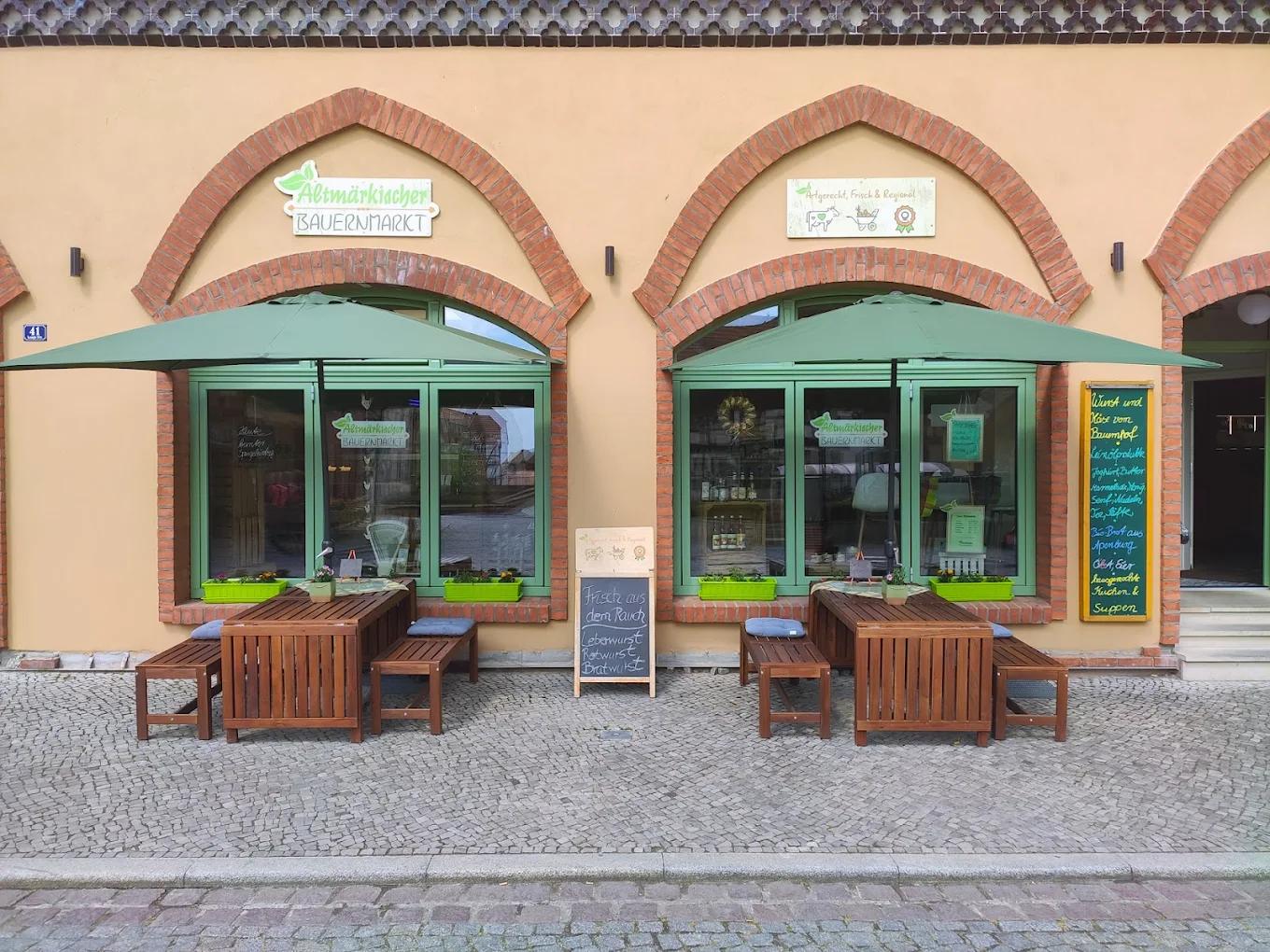 Der Altmärkische Bauernmarkt in der Langen Straße 41 in Tangermünde. Grüne Sonnenschirme
vor den Rundbogenfenstern, Sitzbänke auf dem Kopfsteinpflaster, das Ladenschild über dem Fenster.