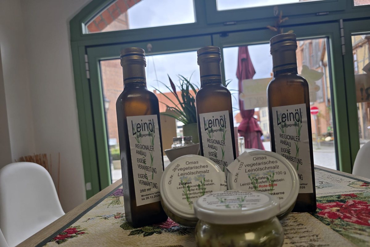 Drei Flaschen kaltgepresstes Leinöl und drei Gläser Omas vegetarisches Leinölschmalz auf einem Tisch am Ladenfenster.