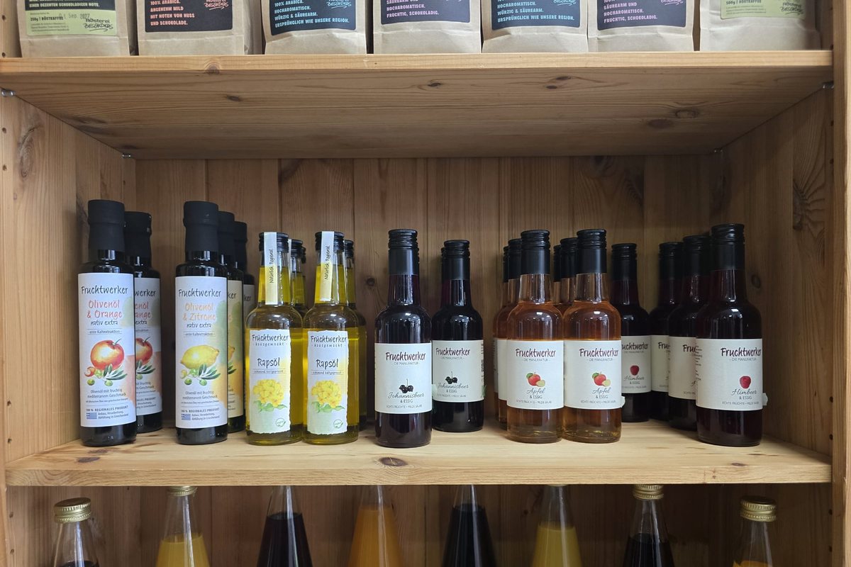 Flaschen von Fruchtwerker im Holzregal: Olivenöl mit Orange und Zitrone, kaltgepresstes Rapsöl und Fruchtessige.