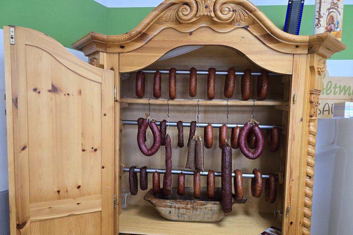 Ein geöffneter Bauernschrank aus hellem Holz, darin hängen Wurstringe und Dauerwürste an Stangen.